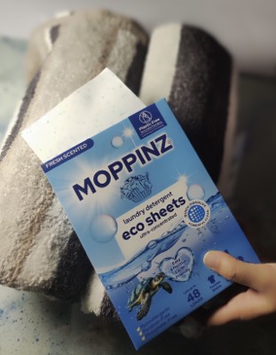 A Moppinz laundry detergent sheets box resting on a stack of freshly folded grey and white towels